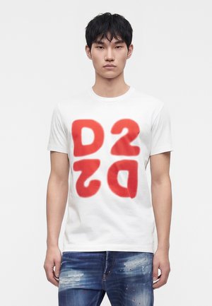 Jonge man met kort zwart haar, een wit t-shirt met grote rode letters "D2" en vervaagde blauwe spijkerbroek met verfspatten.