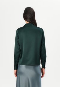 Blusa in raso verde scuro con colletto, maniche lunghe e polsini abbottonati; texture liscia e design corto in vita.