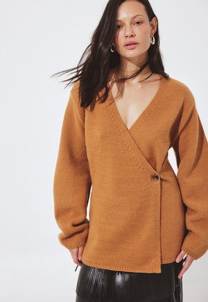 Žena s dlhými tmavými vlasmi, ktorá má na sebe béžový zavinovací sveter s gombíkom a čiernu plisovanú sukňu, stojaca proti jednoliatemu bielemu pozadiu.