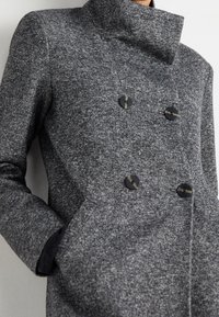 Grå, struktureret dobbeltradet frakke med høj krave, der har to sorte knapper og lommer, lavet af et uld-agtigt materiale.