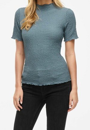 Vrouw draagt een aansluitend, gestructureerd blauw topje met korte mouwen en gerimpelde randen, gecombineerd met een zwarte broek, zichtbaar van schouders tot heupen.