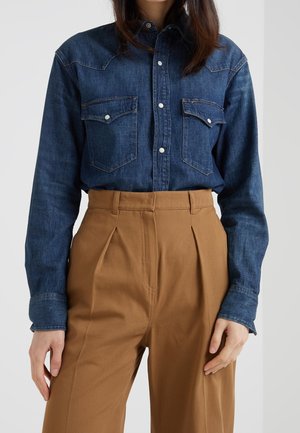 Vrouw die een donkerblauw denim overhemd met knopen draagt, ingestopt in high-waisted camelkleurige geplooide broek, staand tegen een effen achtergrond.