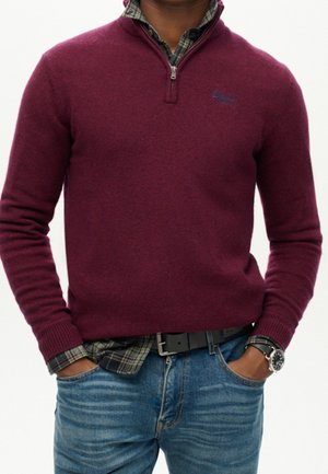 Uomo che indossa un maglione bordeaux con zip a quarto sopra una camicia a scacchi verde, jeans blu, cintura nera e orologio da polso, con le mani nelle tasche.