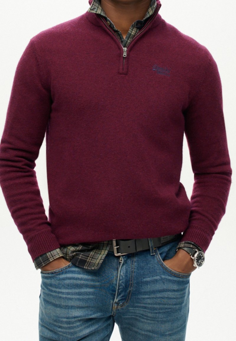 Mann, der einen burgunderfarbenen Viertelreißverschluss-Pullover über einem grünen Karoshemd, blaue Jeans, einen schwarzen Gürtel und eine Armbanduhr trägt, mit den Händen in den Taschen.