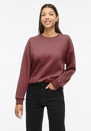 Femeie cu păr lung și întunecat, purtând un pulover bordo cu mâneci lungi și pantaloni negri, zâmbind cu o mână în buzunar.