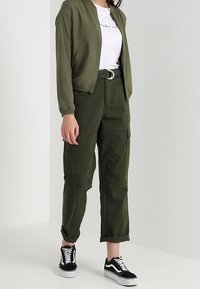 Blouson aviateur vert olive, t-shirt graphique blanc, pantalon cargo vert olive avec poches, et baskets Vans noires. Tissu lisse, coupe décontractée.