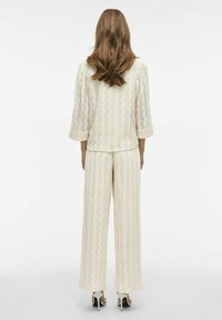 Conjunto de encaje en color crema, con un patrón texturizado de olas. La parte superior tiene mangas de tres cuartos; los pantalones son de pierna ancha, fluyendo hasta el suelo.