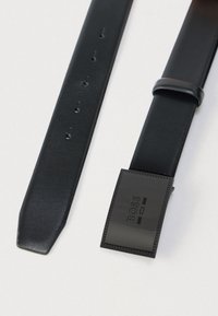 Μαύρη δερμάτινη ζώνη με ορθογώνια μαύρη αγκράφα που φέρει τη λέξη «BOSS», παρουσιασμένη επίπεδη σε ανοιχτόχρωμη επιφάνεια.