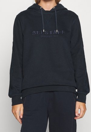 Persoon draagt een zwarte capuchontrui met "BELSTAFF ENGLAND 1924" geborduurd op de borst en bijpassende zwarte broek.