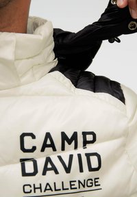 Close-up of a person wearing a white and black puffer jacket with "CAMP DAVID CHALLENGE" printed on the back.