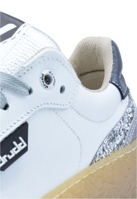 Białe sneakersy z metaliczno-srebrnymi akcentami, teksturowanymi szarymi sznurówkami oraz brązową podeszwą z błyszczącą wstawką na pięcie.