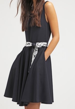 Femme portant une robe noire sans manches avec une jupe évasée et une ceinture en dentelle blanche, mains dans les poches, sur un fond clair.