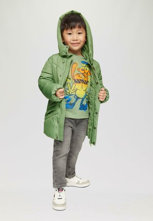 Grüne gepolsterte Jacke mit Kapuze, getragen über einem hellgrünen Shirt mit einem Cartoon-Charakter, kombiniert mit grauen Hosen und weißen Turnschuhen.