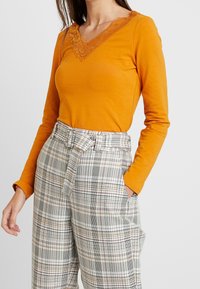 Mustard long-sleeve top with a V-neck adorned with intricate lace, paired with high-waisted plaid trousers featuring a tie belt.