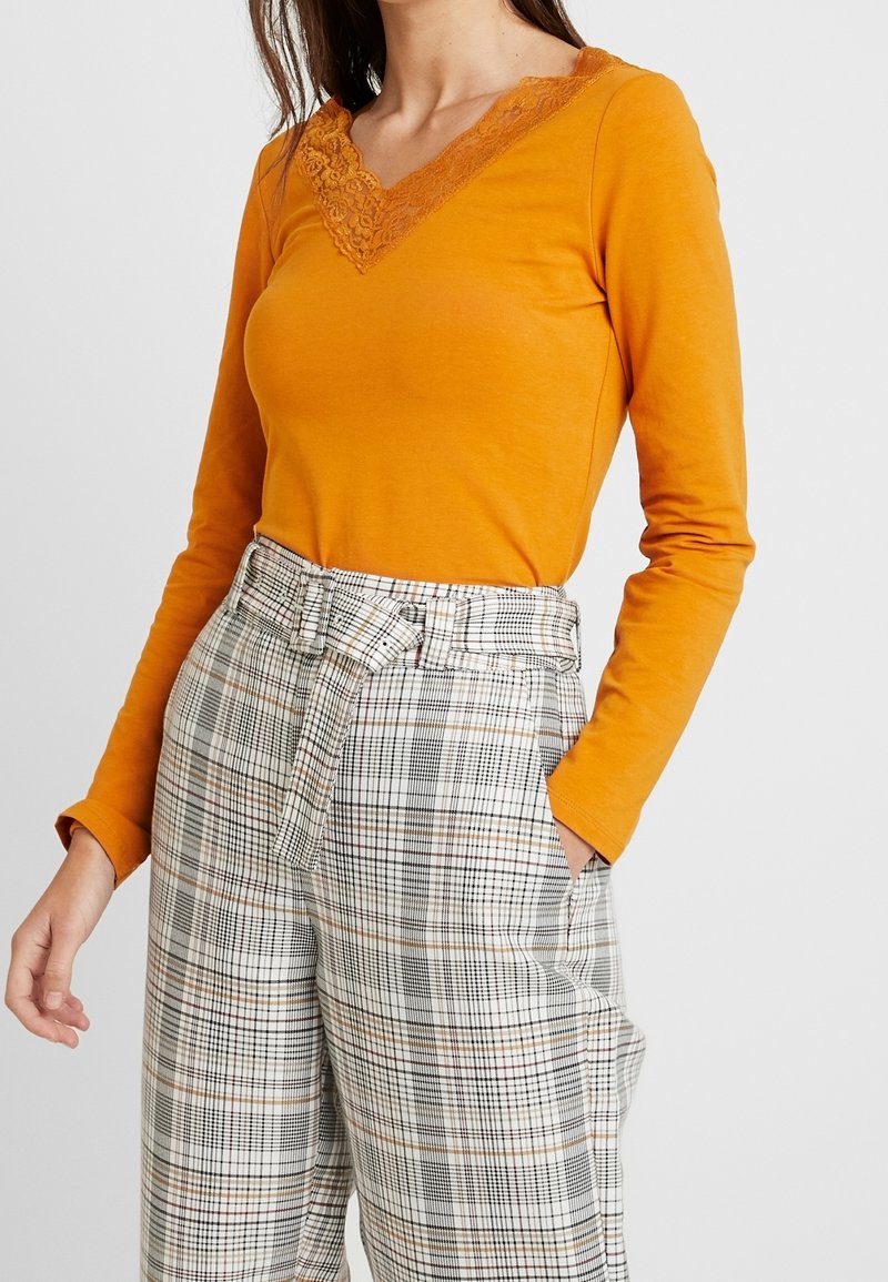Mustard long-sleeve top with a V-neck adorned with intricate lace, paired with high-waisted plaid trousers featuring a tie belt.