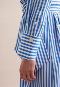 Camicia a righe blu e bianche con polsini abbottonati, decorata con un grande bottone e un piccolo ricamo nero sulla manica.