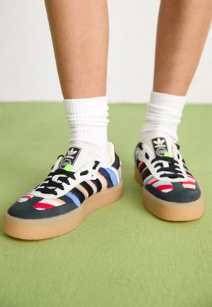 Osoba w białych skarpetkach i kolorowych sneakersach Adidas z gumową podeszwą, stojąca na zielonym dywanie.