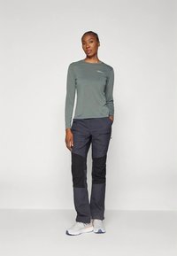 Tamsiai žalia ilgomis rankovėmis palaidinė su apvalia apykakle ir logotipu, derinama su tamsiai pilkomis kelnėmis su juodais akcentais, dėvima su baltais sportbačiais.