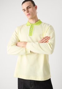 Polo de manga larga en algodón amarillo claro, con un color de cuello verde brillante, tapeta de tres botones y pequeño logotipo bordado.