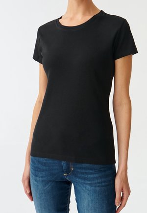 Kvinde iført en tætsiddende sort t-shirt med korte ærmer og rund hals, kombineret med blå denimjeans mod en ensfarvet baggrund.