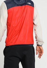 Veste à capuche rouge et grise avec une partie supérieure bleu marine. Elle présente une texture lisse et brillante, des poignets élastiques, et un logo au dos.