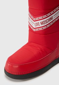 Piros párnázott csizma sima szintetikus felsőrésszel, fekete talppal és egy figyelemre méltó fehér szalaggal, amelyen a "Love Moschino" felirat és szív mintázat található.