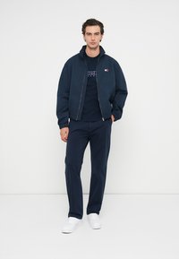 Giacca bomber blu navy con collo alto sopra una t-shirt blu navy, abbinata a pantaloni coordinati e sneakers bianche. Design semplice e pulito.