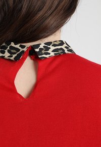 Haut rouge avec un col à imprimé léopard, doté d'une ouverture en forme de goutte d'eau dans le dos et d'une fermeture par bouton. Le tissu est lisse et ajusté.