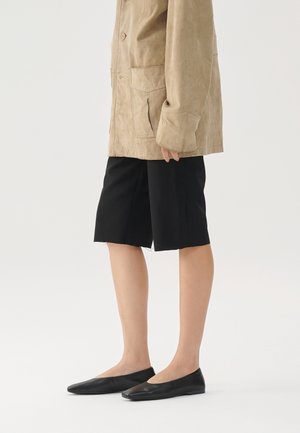 Person iført en beige jakke, sorte knælange shorts og sorte flade sko mod en ensartet lys baggrund.