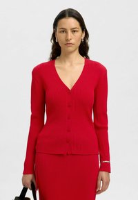 Röd räfflad cardigan med V-ringning, långa ärmar och fem framknappar, tillsammans med en matchande midikjol. Slät textur.