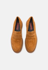 Mocasines de ante marrón con un diseño de mocasín, que cuentan con una correa decorativa, suela de goma y plantilla negra con un logo en rojo.