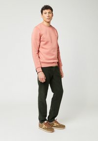 Jonge man die staat, gekleed in een roze sweatshirt, donkergroene corduroy broek en bruine sneakers met oranje veters, tegen een effen lichte achtergrond.