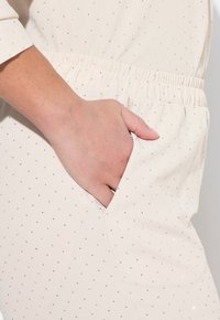 Beige tyg med små silverpaljetter, utrustad med resårmidja och sidoficka, där en hand vilar nära ficköppningen.