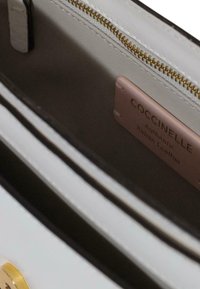 Interiør af hvid lædertaske med guldfarvet lynlås, lysebeige foer og en lyserød label med teksten "Coccinelle Authentic Italian Leather."