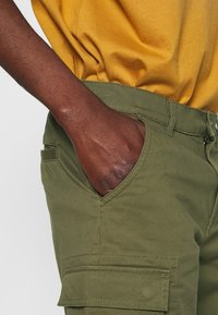 Pantalons cargo vert olive avec une poche latérale et une fermeture éclair, montrés avec une main dans la poche. Fabriqués en tissu robuste et avec des lignes épurées.