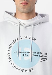 Sudadera gris claro con un gráfico circular en la parte frontal en texto azul y negro. Tiene una capucha con cordón y una textura de tela suave.