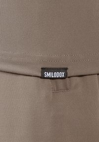 Brązowa tkanina tekstylna o gładkiej fakturze, z czarną naszywką "SMILODOX" przyszytą do szwu.