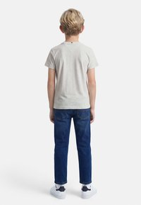 Αγόρι με κοντά ξανθά μαλλιά στέκεται πλάτη, φοράει ανοιχτό γκρι κοντομάνικη μπλούζα, μπλε τζιν και λευκά αθλητικά παπούτσια με μαύρες λεπτομέρειες.