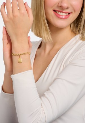 Smiling woman with short blonde hair wearing a white top, showing a gold chain bracelet with a rectangular zodiac Virgo charm on her wrist.