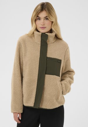 Femme portant une veste en polaire sherpa beige avec un col montant, une poche poitrine zippée vert olive et une patte de boutonnage à l'avant, posant devant un fond uni.