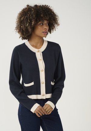 Navy cardigan with cream accents, featuring four buttons, two front pockets, and a ribbed collar and cuffs. Soft, textured knit fabric.
