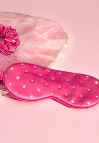 Mascherina per gli occhi in satin rosa con texture imbottita e piccole borchie argentate, accompagnata da un elastico coordinato rosa. Entrambi gli oggetti su uno sfondo chiaro.
