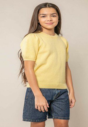 Meisje met lang bruin haar, gekleed in een geel truitje met korte mouwen en een spijkershort, tegen een beige achtergrond.