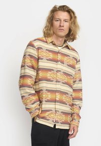 Man met schouderlang blond haar, gekleed in een gele, oranje en rode geblokte overhemd en zwarte broek, staand met zijn handen in zijn zakken.
