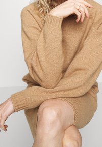 Persoon in een beige gebreide jurk zit met gekruiste benen, steunt met de kin op de hand, heeft krullend haar en een simpele ring om de vinger.