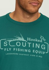 Turkoois katoenen T-shirt met een zeefgedrukte graphic met visserij-thema tekst en afbeeldingen in lichtere tinten, ronde halslijn en korte mouwen.