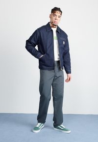 Veste en denim foncé avec coutures contrastées, t-shirt graphique blanc, pantalon gris et baskets vertes. Le mannequin est debout, les mains dans les poches.