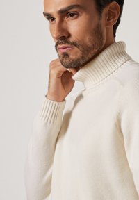 Turtlenecktröja i mjuk krämfärgad tyg, med ribbad krage och manschetter, slät yta och figurnära design. Närbild av modellens profil.