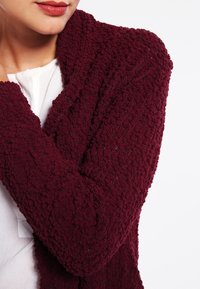 Bordeaux gebreide cardigan met een gestructureerde, pluche stof en een open voorkant ontwerp. Kenmerken zijn een subtiel patroon en een gezellige sjaalkraag.