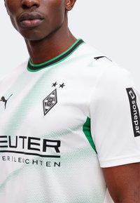 Közeli kép egy személyről, aki fehér és zöld focimezt visel, amelyen a Borussia Mönchengladbach logója, a Puma emblémája és a Sonepar szponzor látható az ujján.